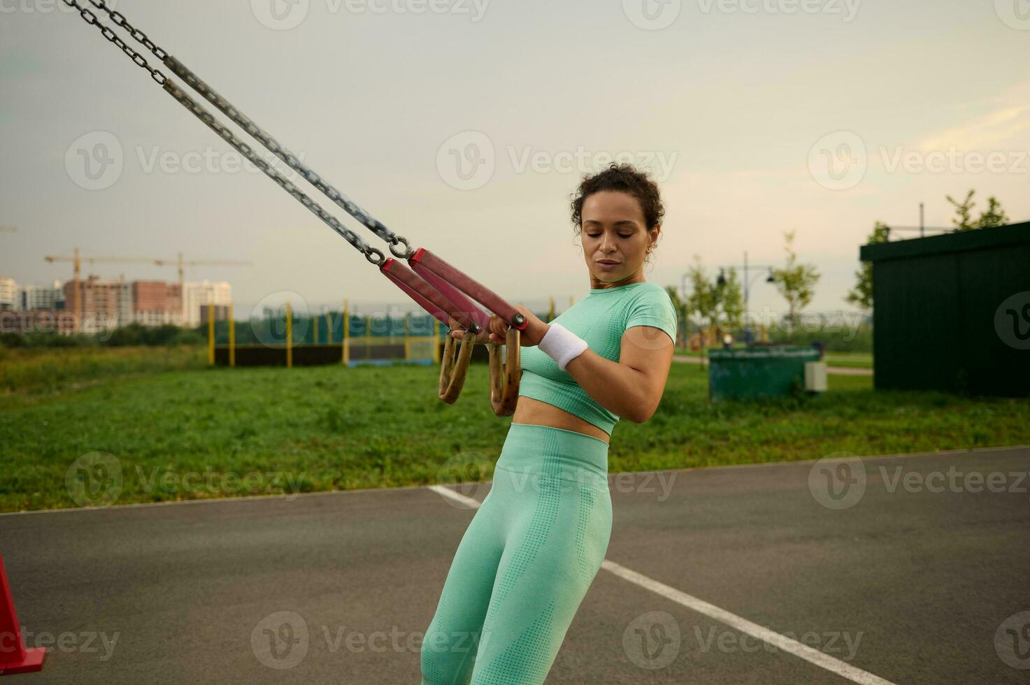 Side view of a sporty determinate middle aged African American, mixed race woman doing arm exercises with suspension straps while performing an outdoor cross training in the sports field photo