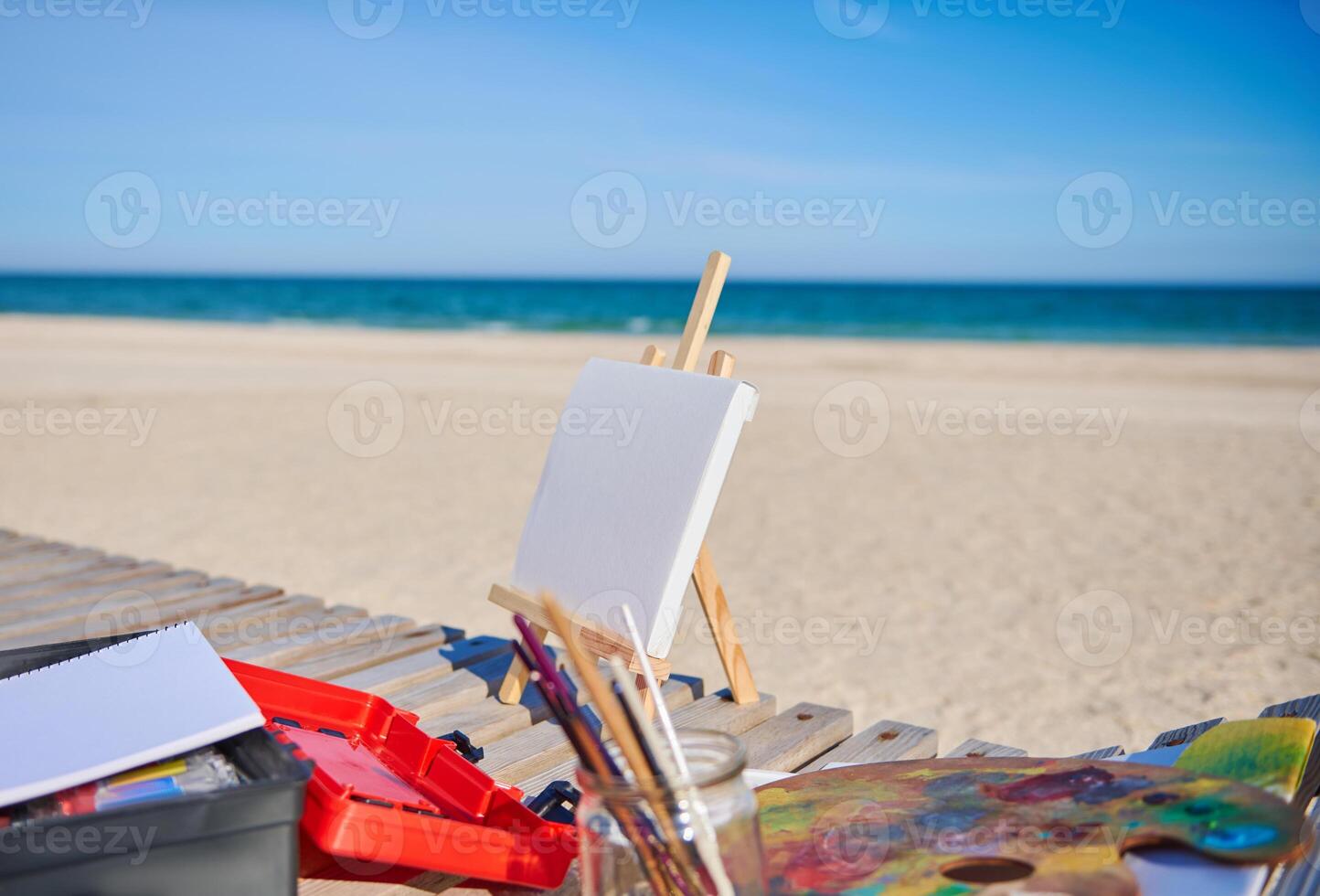White blank canvas on a wooden table easel next to paints and brushes on a wooden deck chair on the beach against the beautiful blue sea background photo