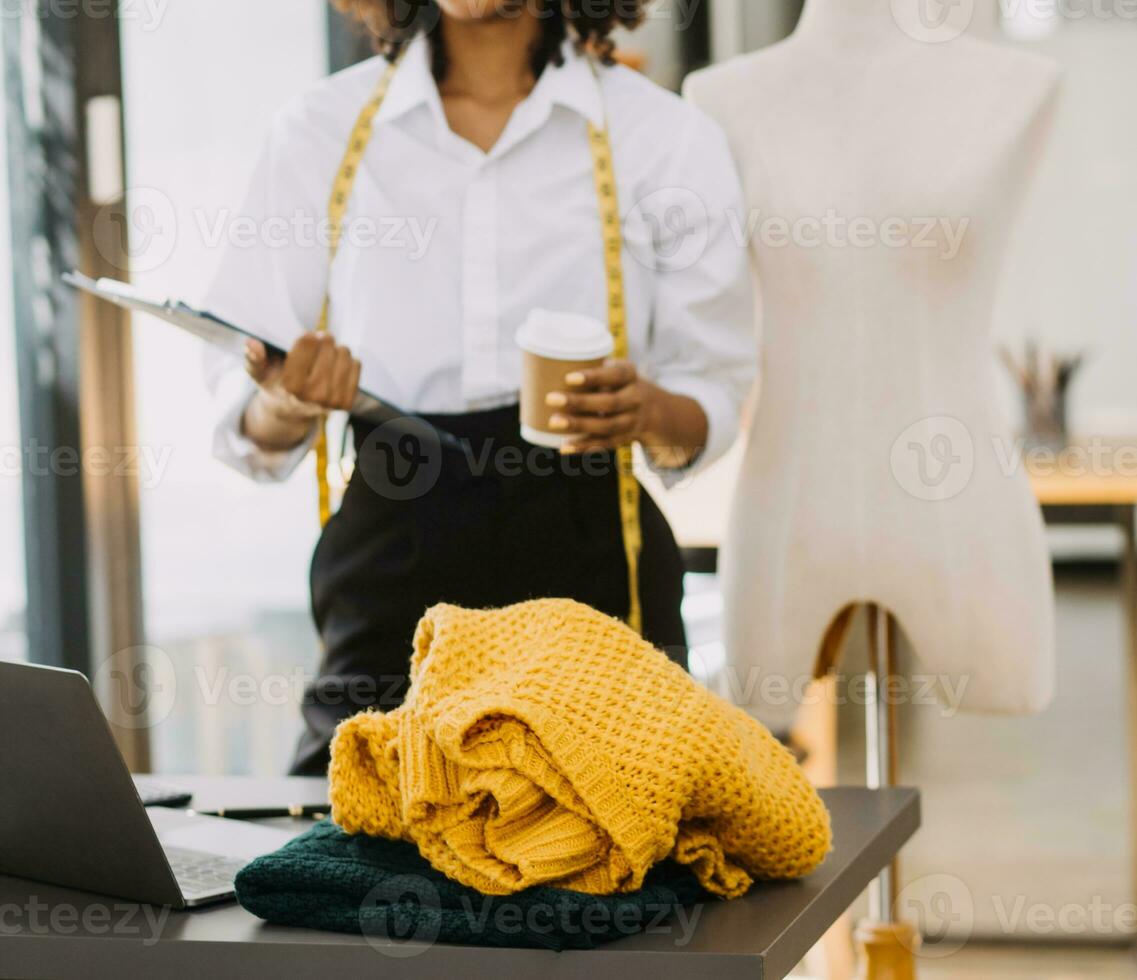 Female fashion, designer, Looking at Drawings and Sketches that are Pinned to the Wall Behind Her Desk. Studio is Sunny. Personal Computer, Colorful Fabrics, Sewing Items are Visible. photo