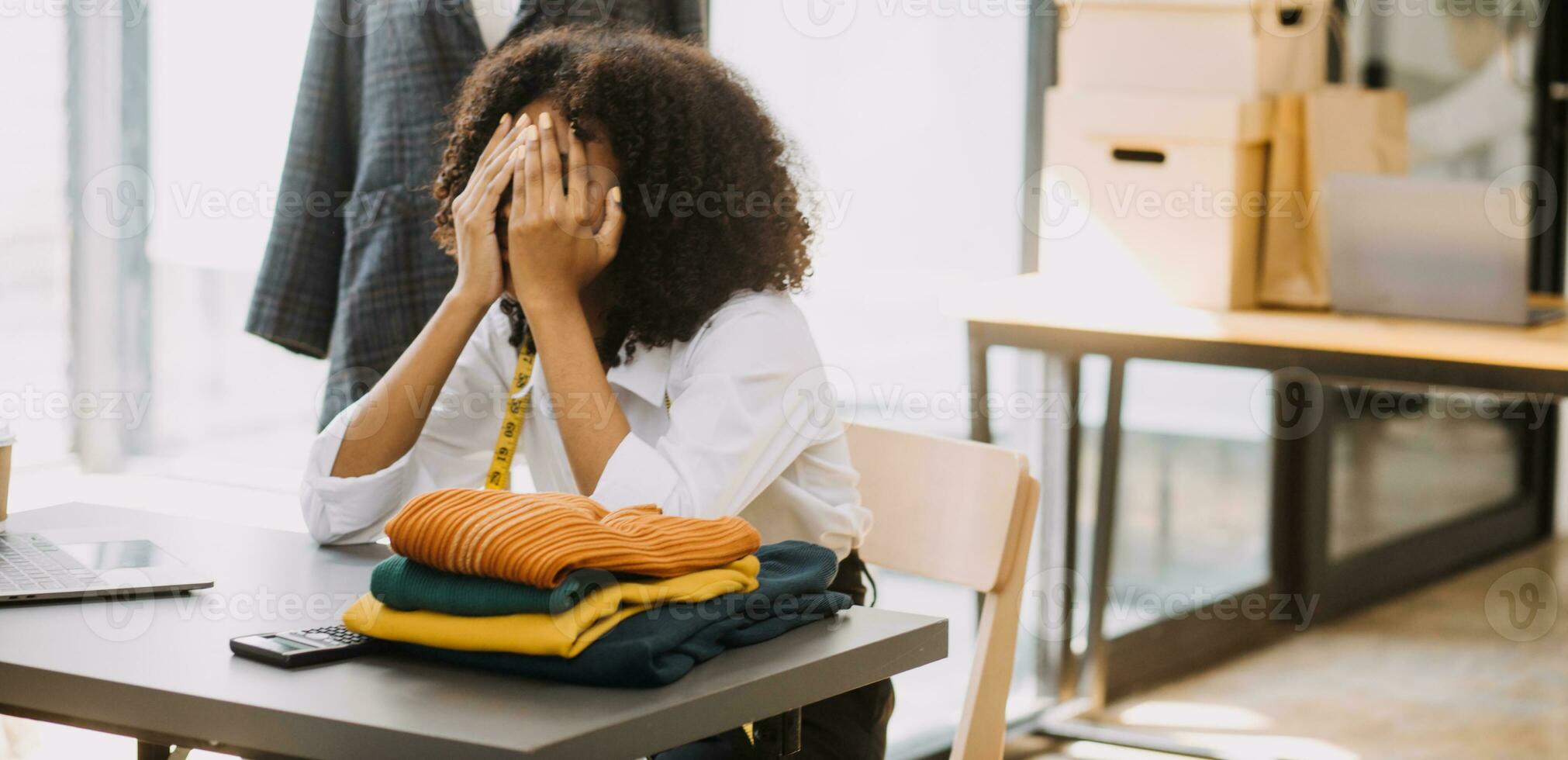 Female fashion, designer, Looking at Drawings and Sketches that are Pinned to the Wall Behind Her Desk. Studio is Sunny. Personal Computer, Colorful Fabrics, Sewing Items are Visible. photo