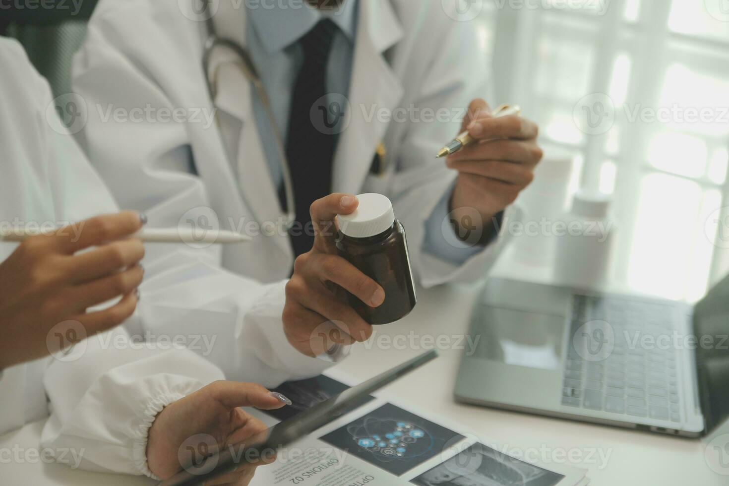 Confident doctor man holding a pill bottle and writing while talking with senior patient and reviewing his medication at office room. photo