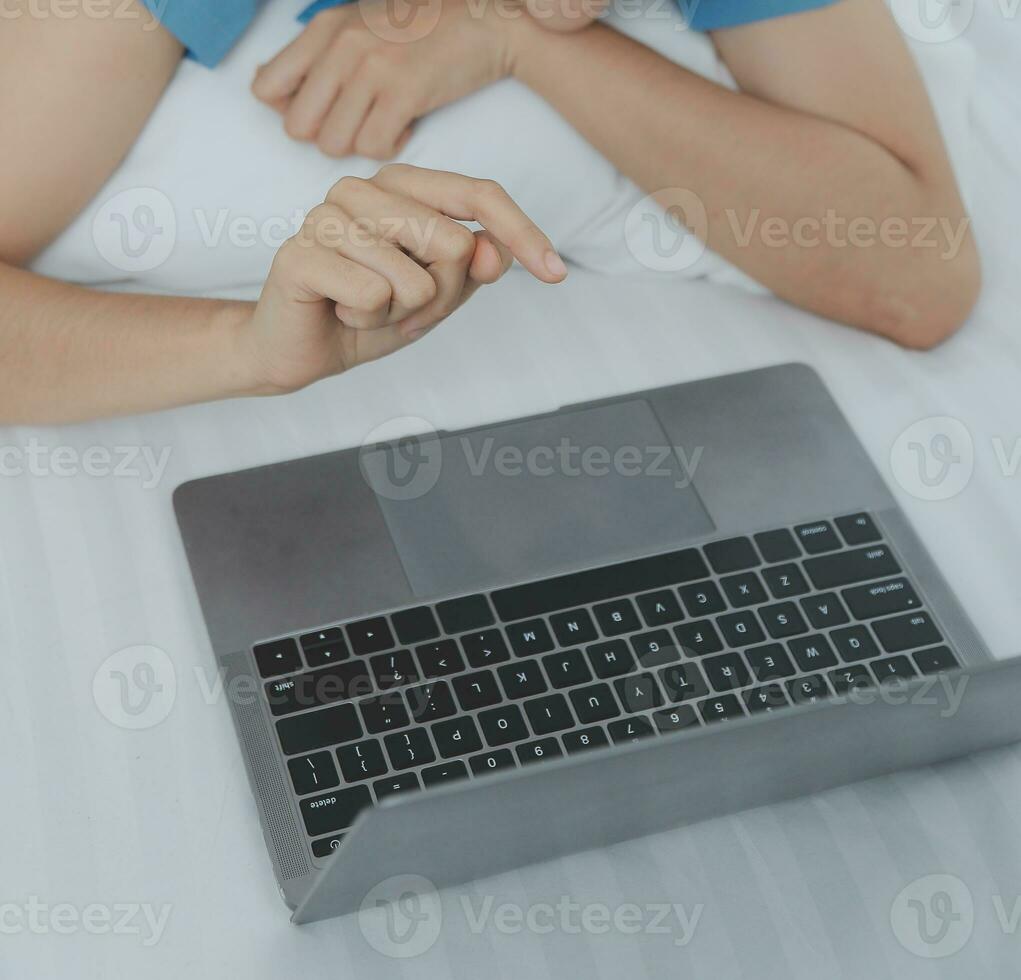 Young Asian woman with beautiful face, long hair, blue shirt lying on bed in white bedroom at home with cup of coffee and video call with laptop talking to relatives on vacation. holiday concept. photo