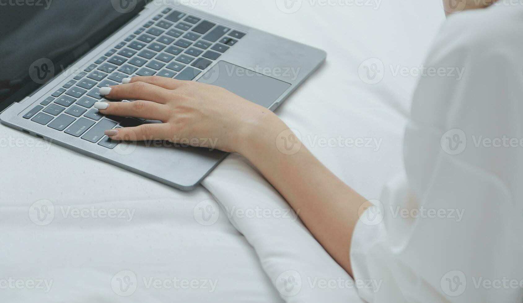 Happy young female in casual clothes smiling and looking at camera while using laptop on bed in bedroom at home photo