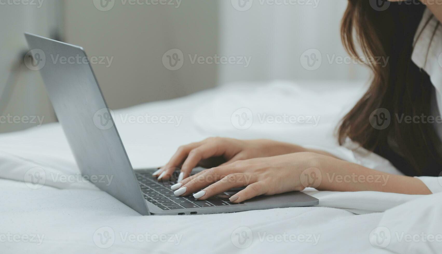 Happy young female in casual clothes smiling and looking at camera while using laptop on bed in bedroom at home photo