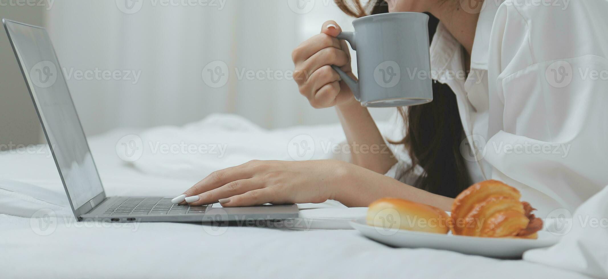 Happy young female in casual clothes smiling and looking at camera while using laptop on bed in bedroom at home photo