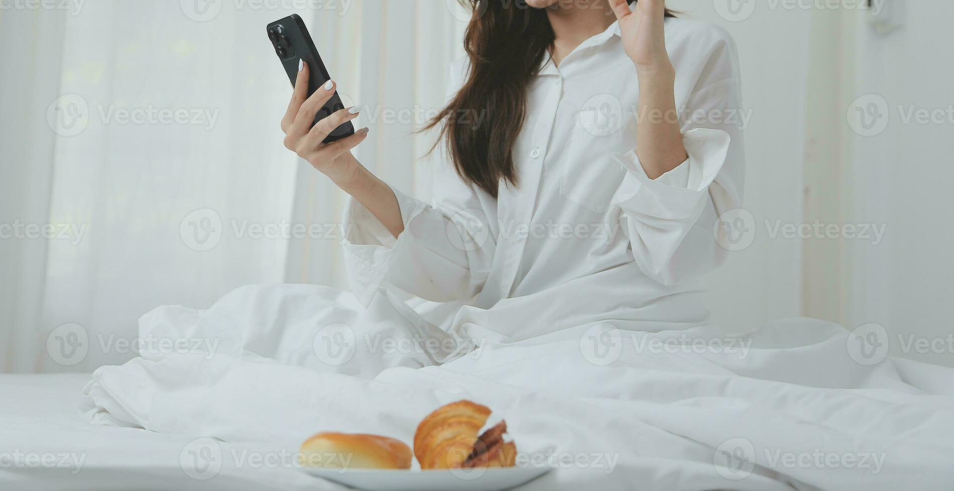 Happy young female in casual clothes smiling and looking at camera while using laptop on bed in bedroom at home photo