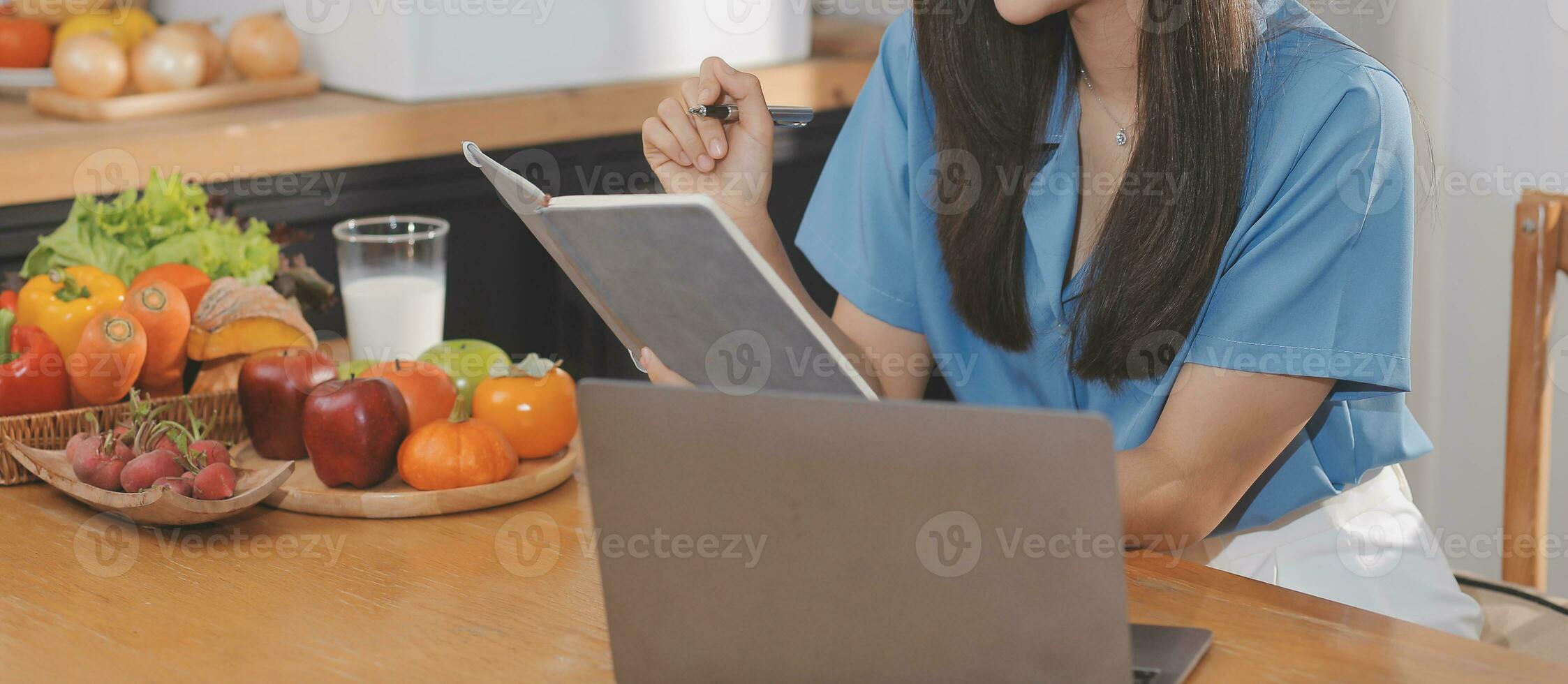 A young woman with a beautiful face in a blue shirt with long hair eating fruit sitting inside the kitchen at home with a laptop and notebook for relaxation, Concept Vacation. photo