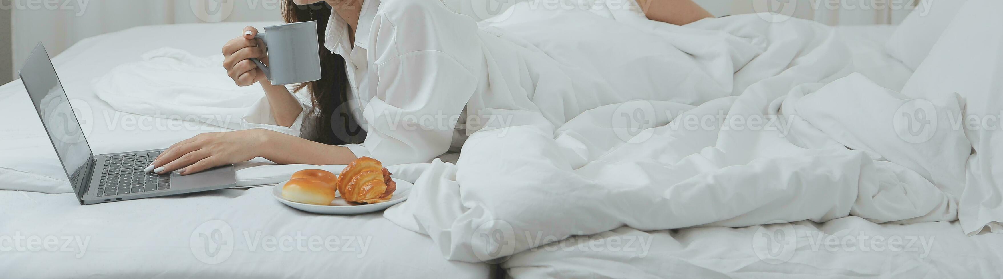 Happy young female in casual clothes smiling and looking at camera while using laptop on bed in bedroom at home photo