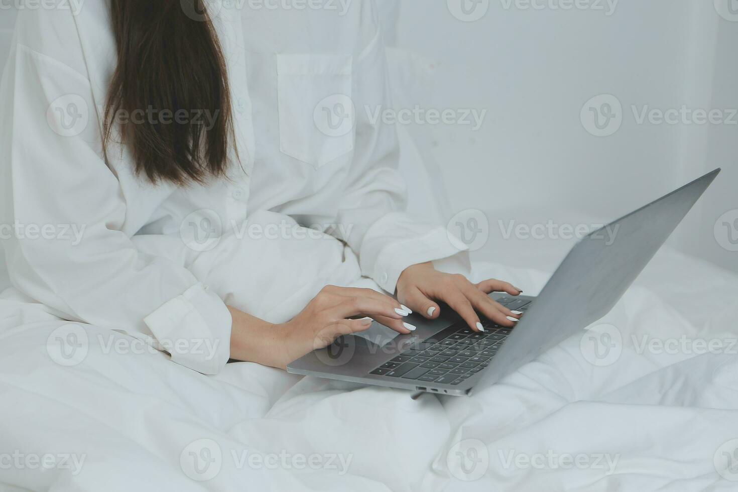Happy young female in casual clothes smiling and looking at camera while using laptop on bed in bedroom at home photo