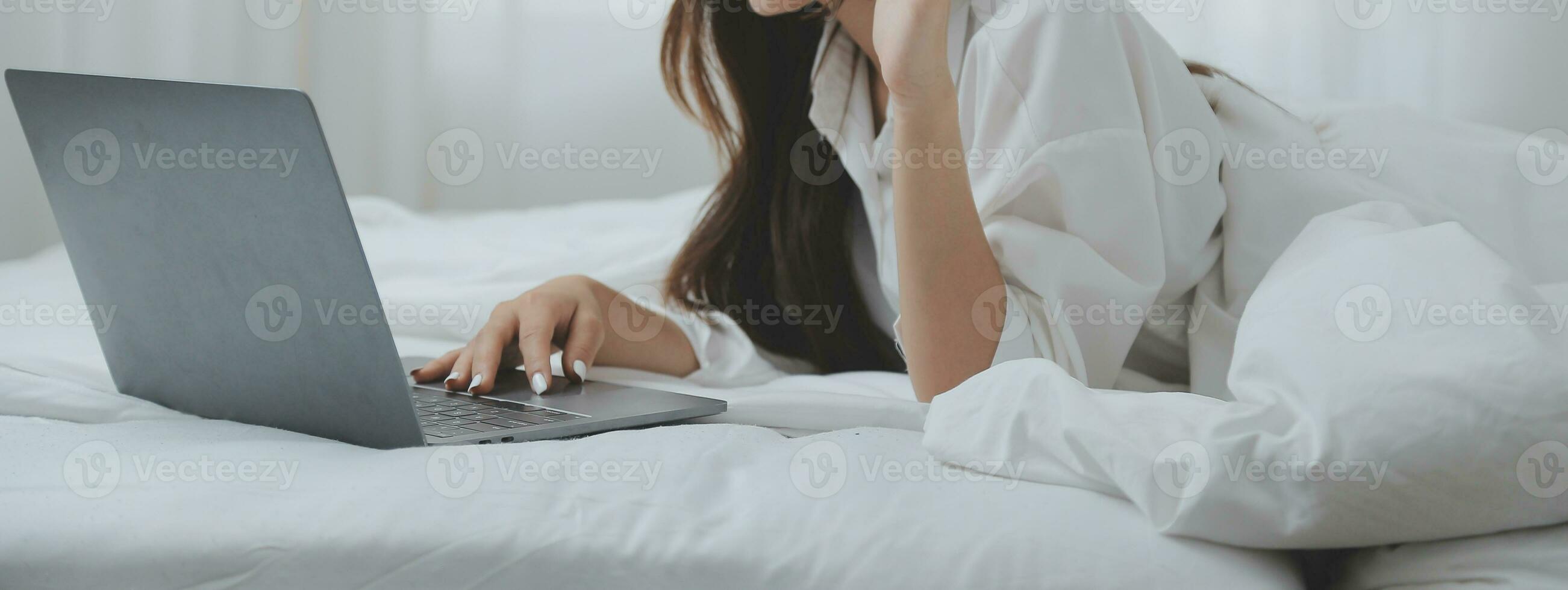 Happy young female in casual clothes smiling and looking at camera while using laptop on bed in bedroom at home photo