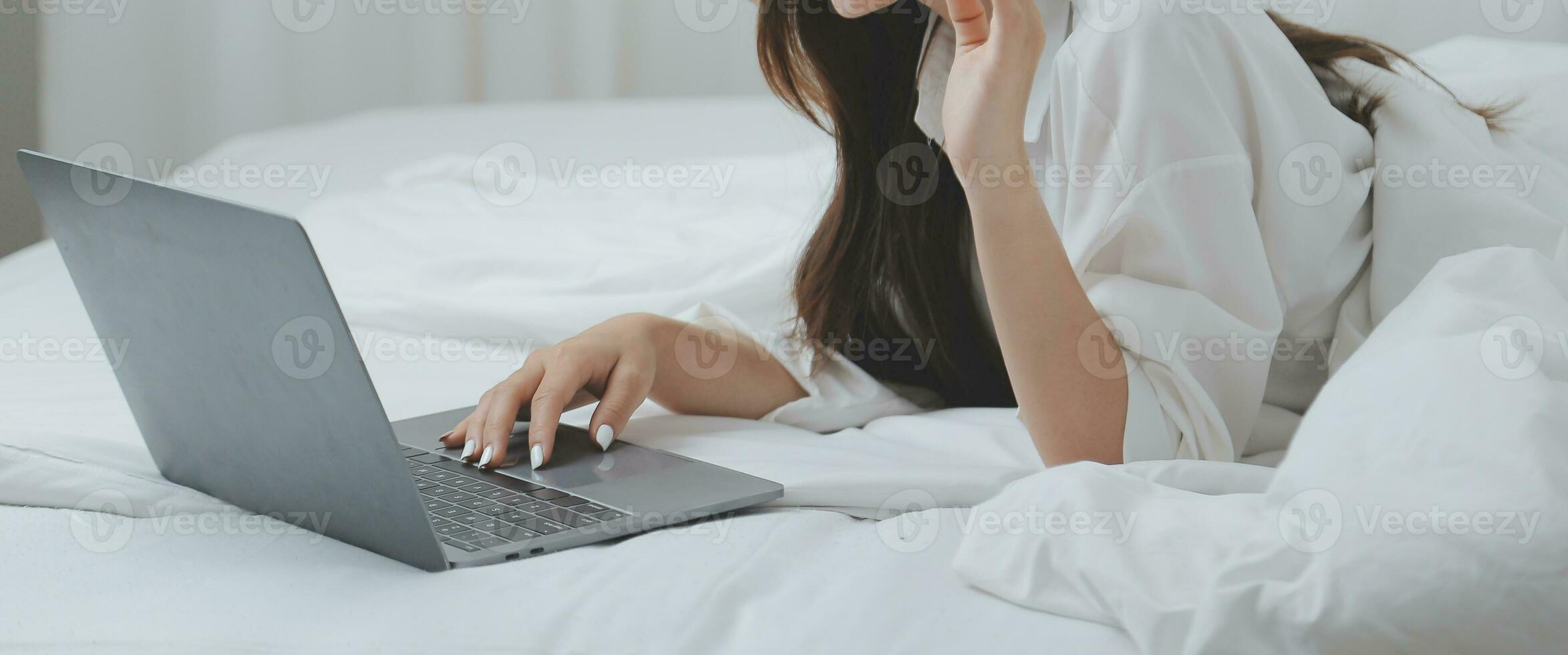 Happy young female in casual clothes smiling and looking at camera while using laptop on bed in bedroom at home photo