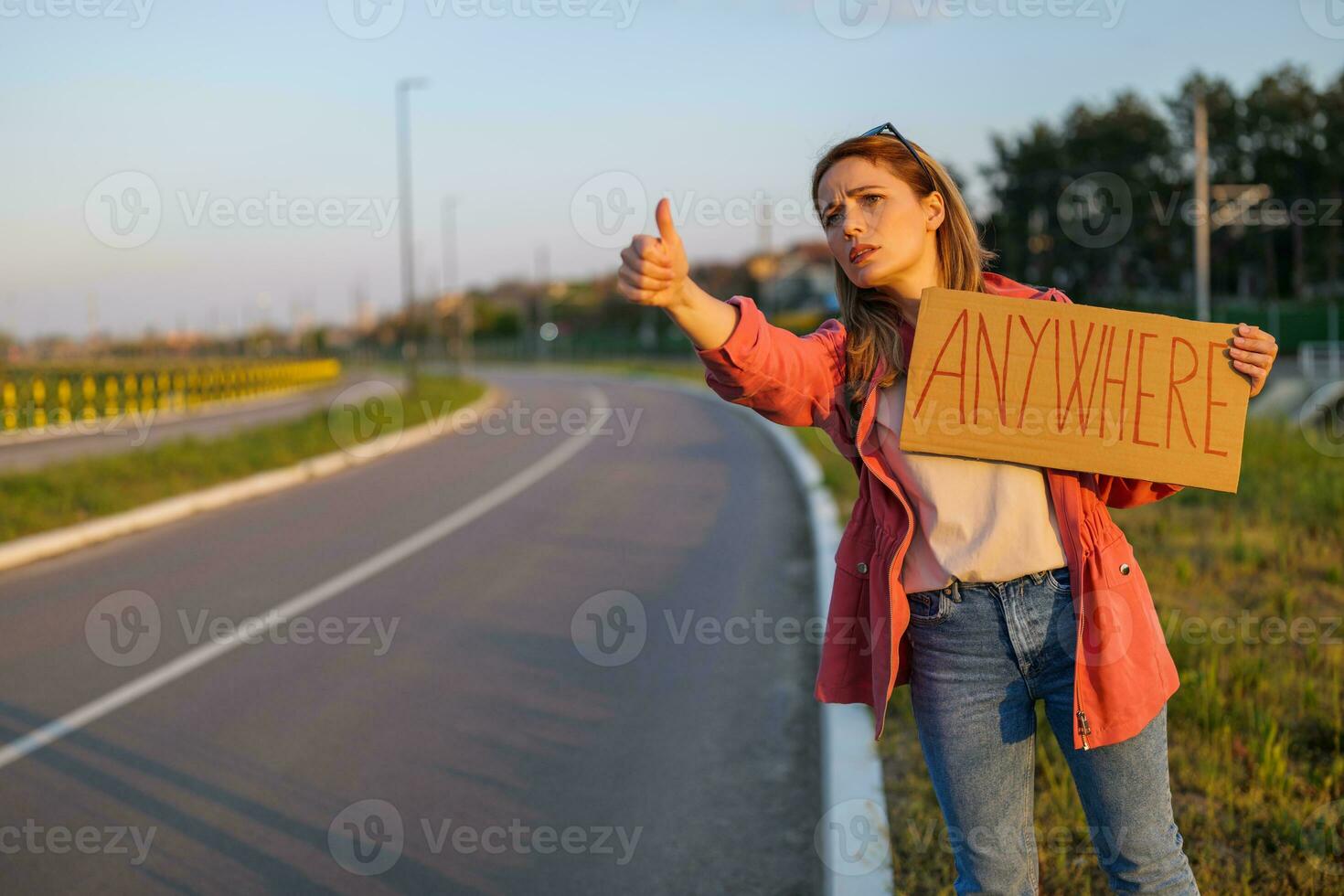 Woman is hitchhiking on roadside trying to stop car. She is holding cardboard with inscription. photo