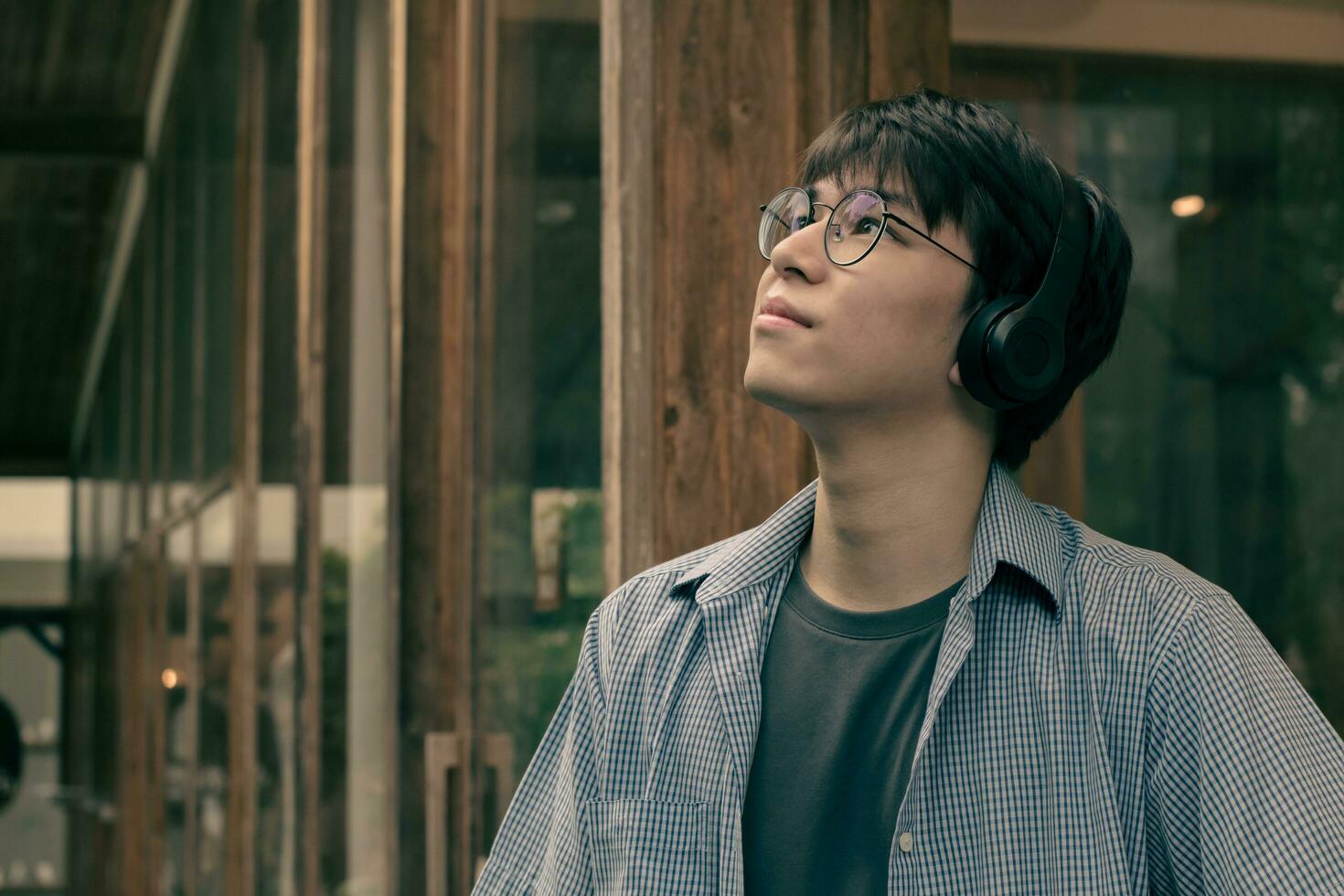 Portrait of asian young adult in blue shirt, wearing eyeglasses, holding earphones and standing in front of the door of coffee shop, concept for lifestyle of teenagers around the world. photo