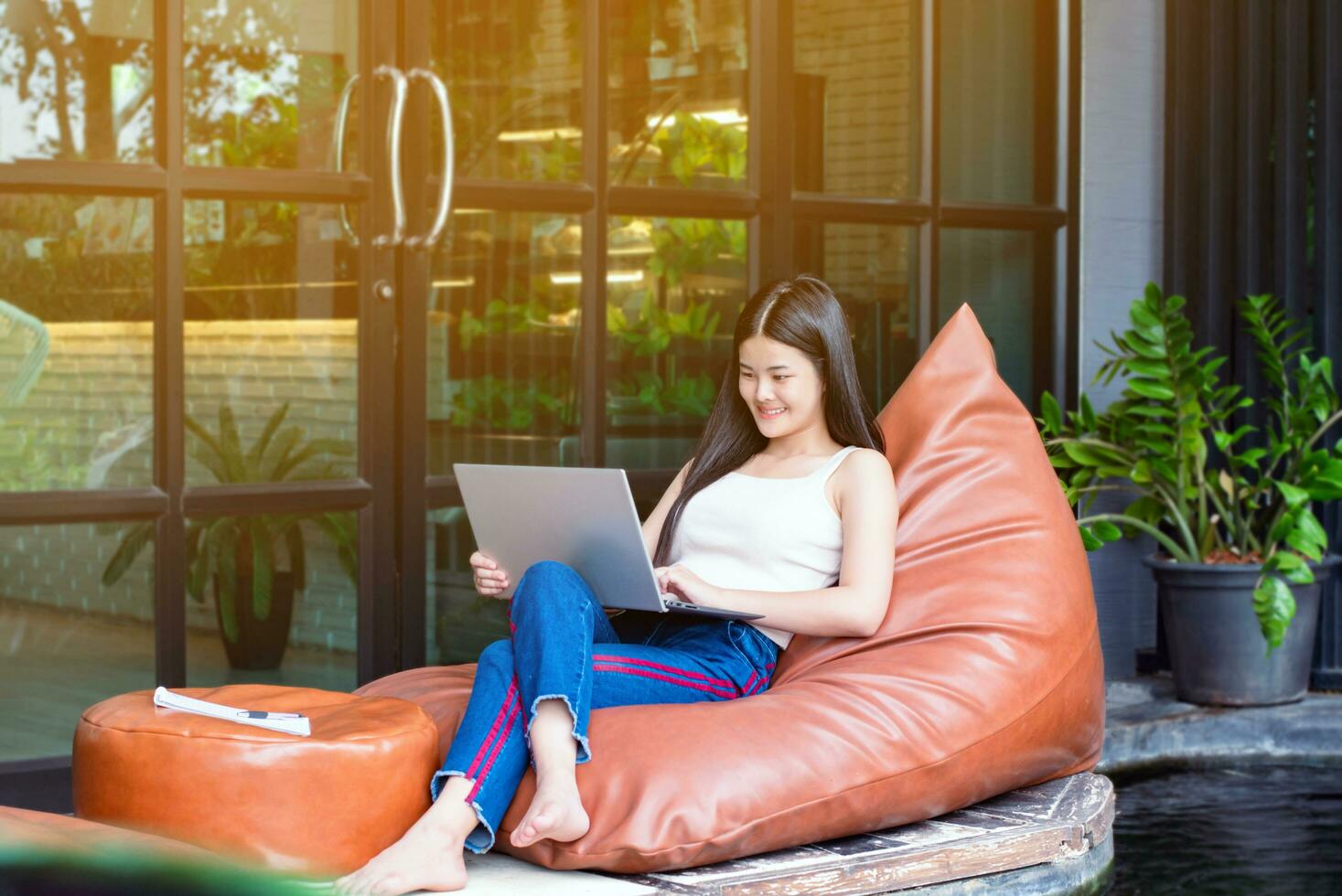 Young adult asian businesswoman sitting on pillow, holding pen and shortnote, looking down to her laptop in front of glassdoor of the cafe during her long vacation, digital nomad of woman concept. photo