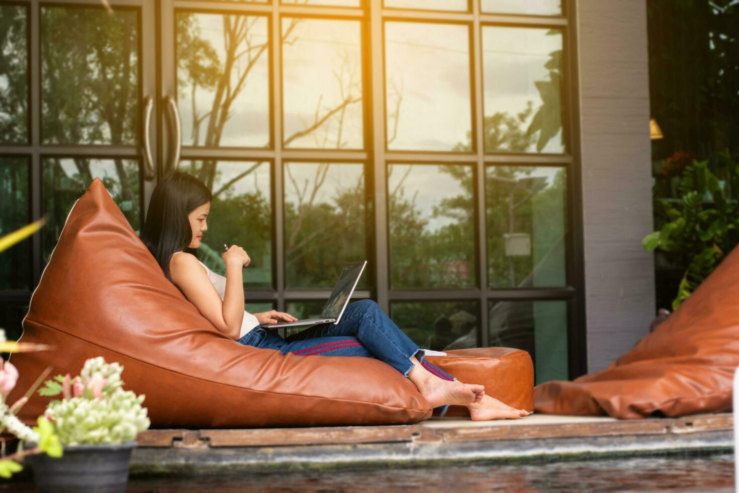 joven adulto asiático mujer de negocios sentado en almohada, participación bolígrafo y nota corta, mirando abajo a su ordenador portátil en frente de puerta de vidrio de el café durante su largo vacaciones, digital nómada de mujer concepto. foto