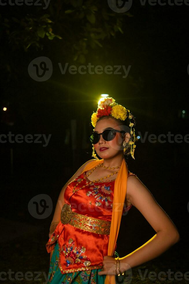 a Sundanese dancer poses in front of a shining light while wearing an orange costume with sunglasses and flowers on her head photo