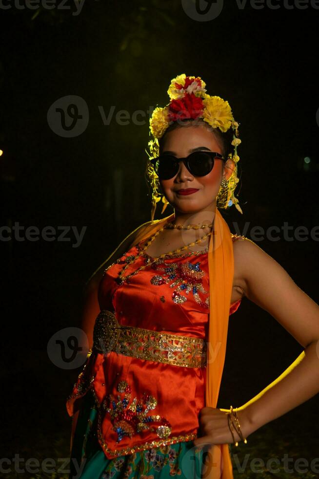 un sundanés bailarín poses en frente de un brillante ligero mientras vistiendo un naranja disfraz con Gafas de sol y flores en su cabeza foto