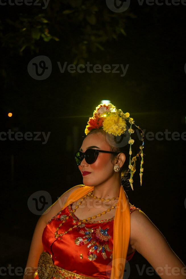 un sundanés bailarín poses en frente de un brillante ligero mientras vistiendo un naranja disfraz con Gafas de sol y flores en su cabeza foto