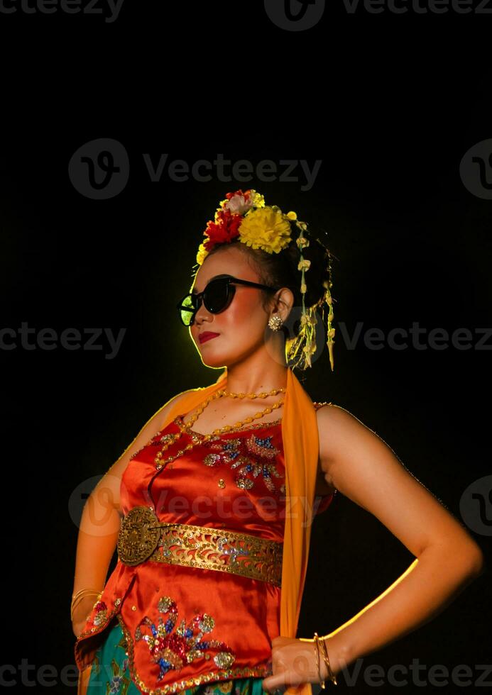 a Sundanese dancer poses in front of a shining light while wearing an orange costume with sunglasses and flowers on her head photo