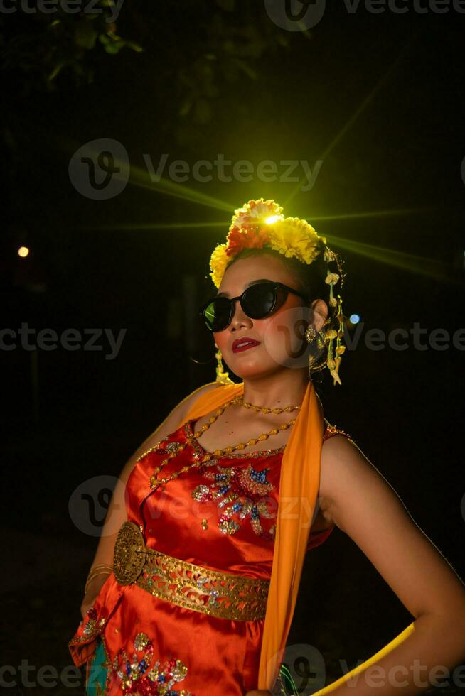 un sundanés bailarín poses en frente de un brillante ligero mientras vistiendo un naranja disfraz con Gafas de sol y flores en su cabeza foto