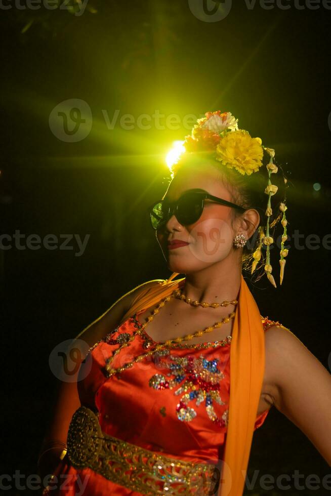 a Sundanese dancer poses in front of a shining light while wearing an orange costume with sunglasses and flowers on her head photo