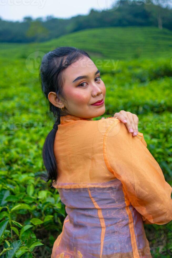 an Asian woman in an orange dress and blue skirt standing among the green tea leaves photo