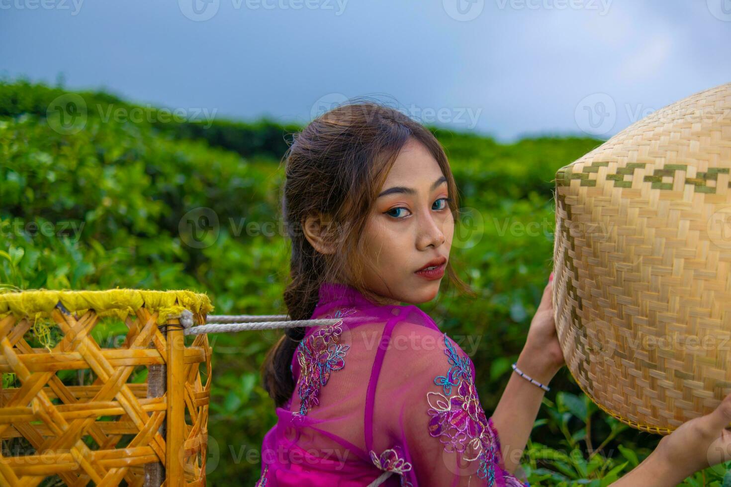 un vietnamita niña es en pie en el medio de un té jardín mientras que lleva un bambú cesta y participación un bambú sombrero foto