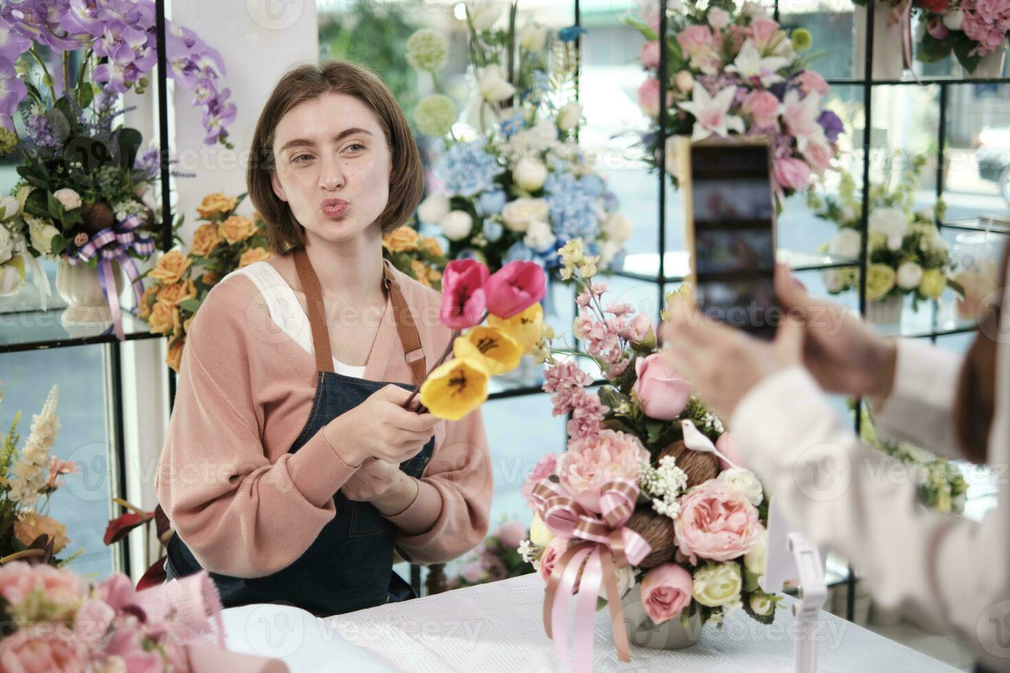 comercio electrónico negocio, uno joven blanco hembra florista demuestra y muestra floral preparativos vía en línea En Vivo transmisión con teléfono inteligente solicitud en brillante flor comercio, hermosa flores almacenar. foto