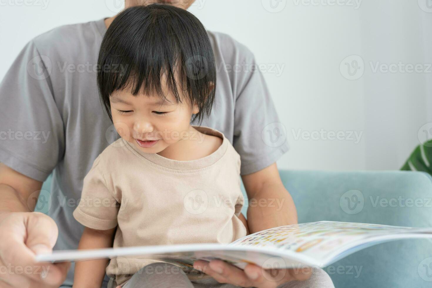 Happy Asian father relax and read book with baby time together at home. parent sit on sofa with daughter and reading a story. learn development, childcare, laughing, education, storytelling, practice. photo