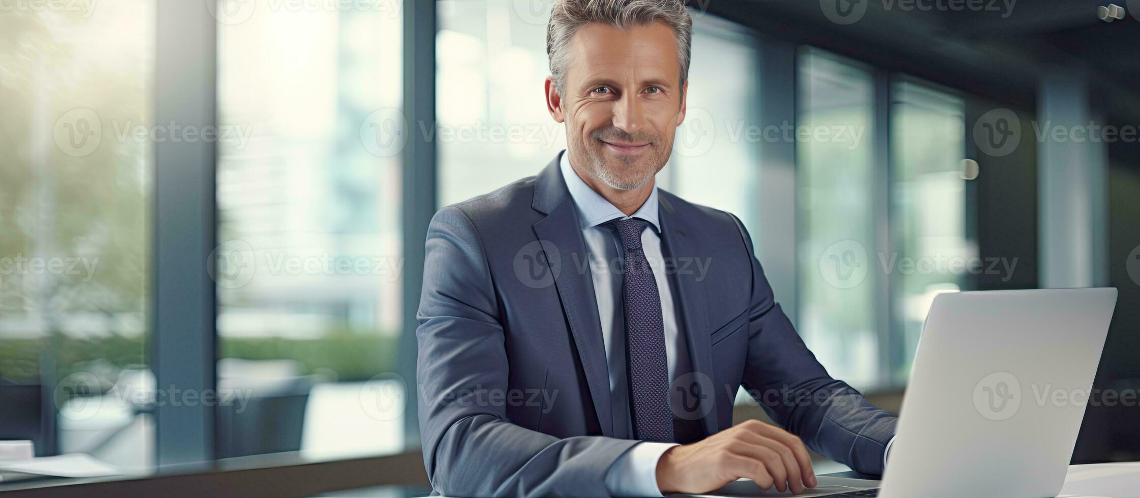 Middle aged professional sitting at desk in office working on laptop and taking notes photo