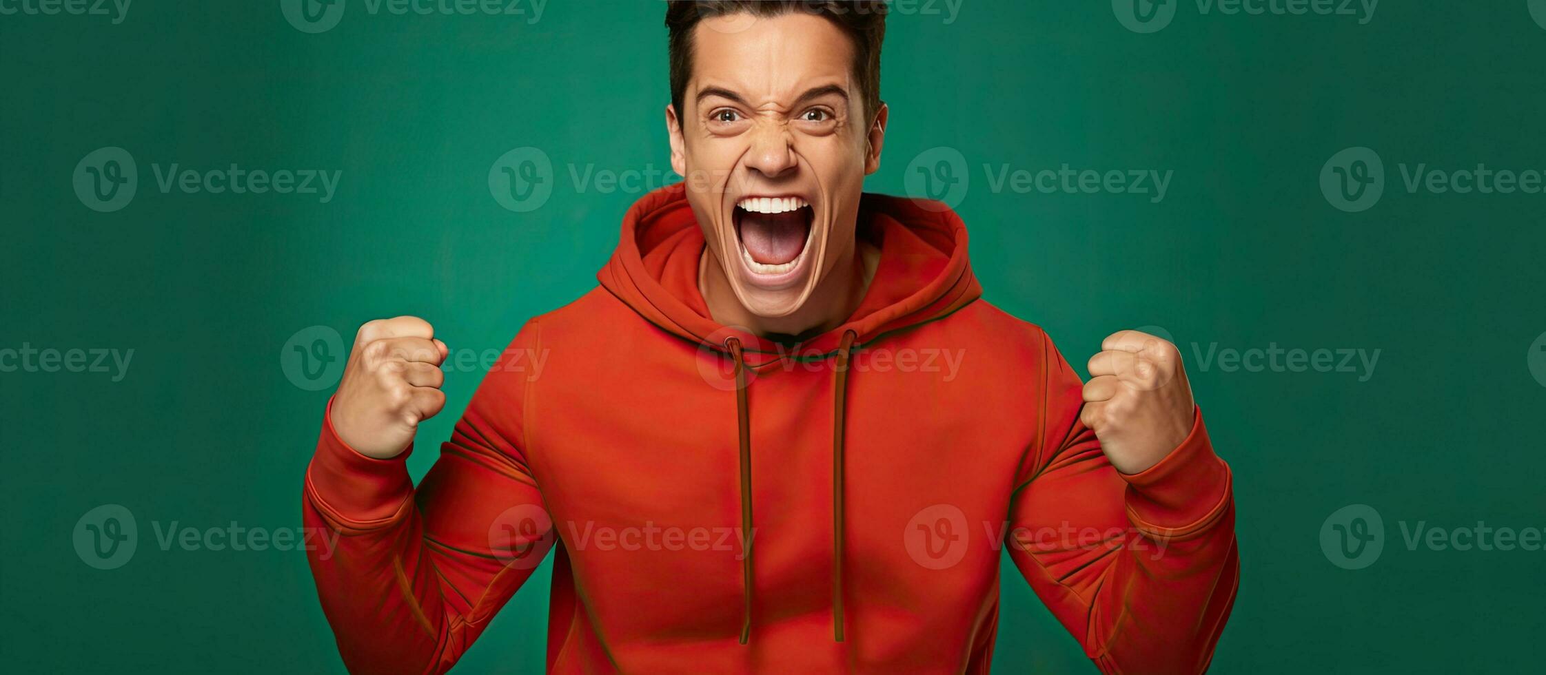 Brunet man in his 20s wearing a red t shirt and green jacket celebrates with a winner gesture isolated on an orange background photo
