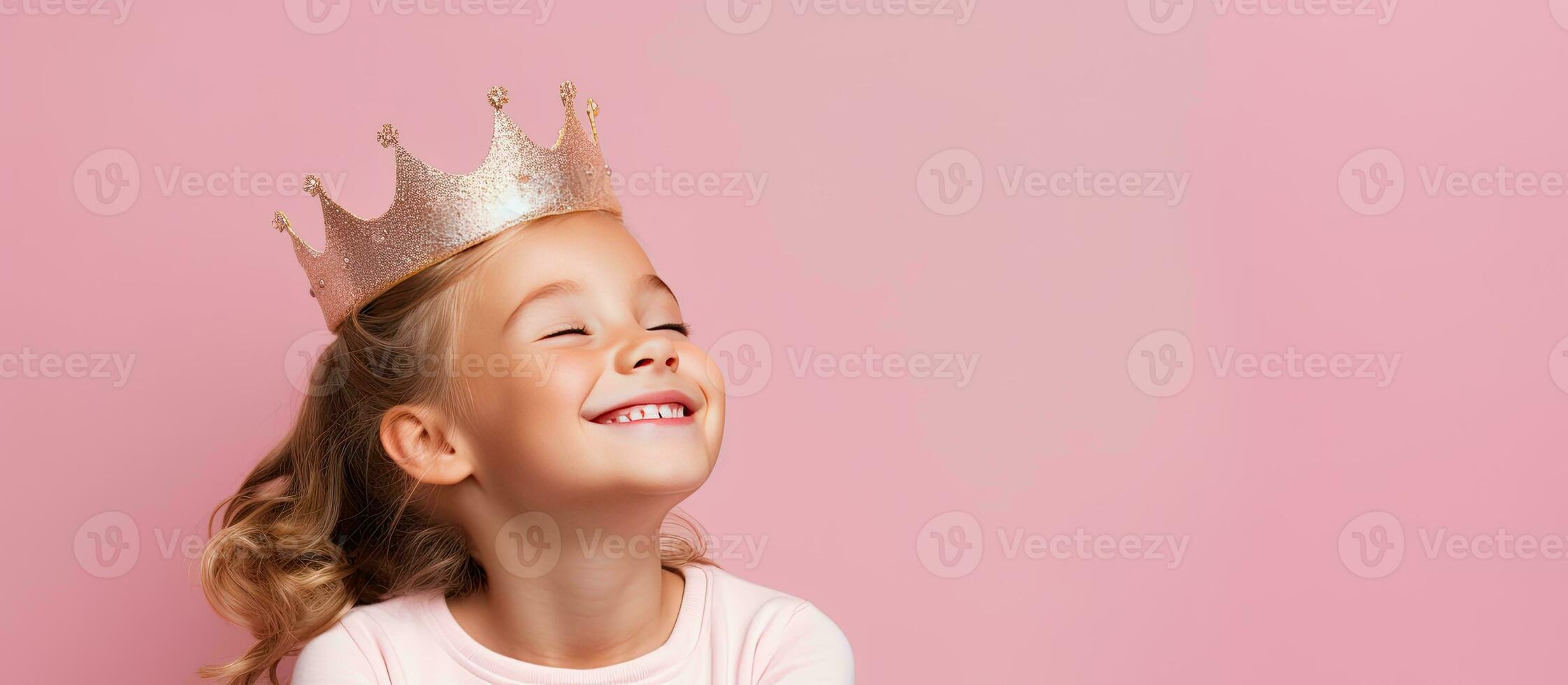 Happy young girl wearing toy crown posing over pink background looking aside with dreamy expression photo