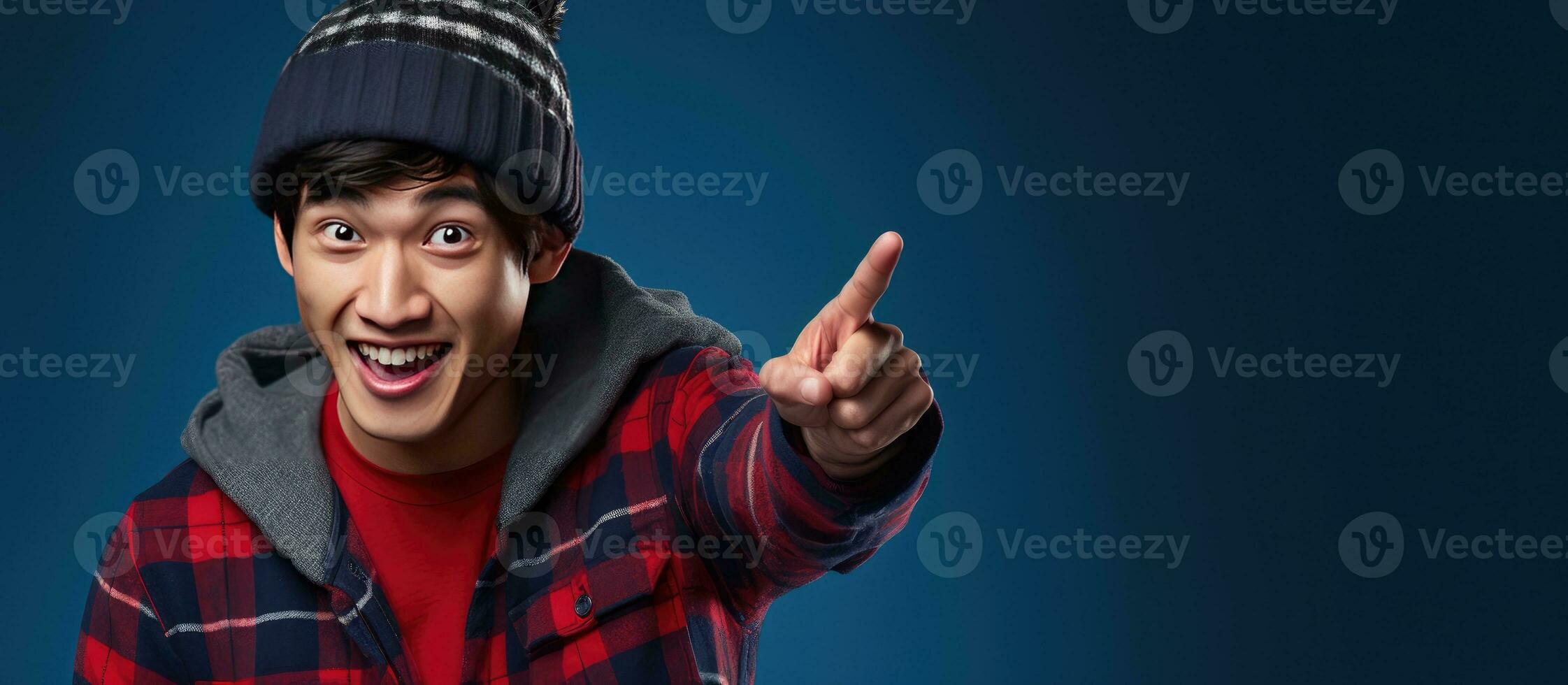 An enthusiastic Asian man with a beanie hat and red plaid flannel shirt talking on his phone and pointing left isolated on blue photo