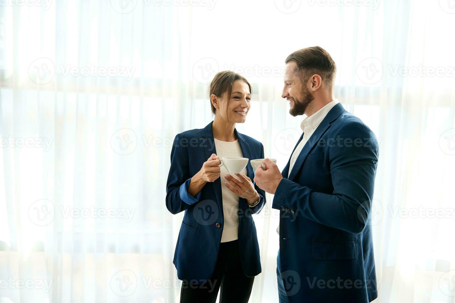 Two pretty colleagues, partners, a pretty woman and a handsome man in a business suit talking together over a cup of coffee, standing next to each other by the window in hotel room at work trip photo