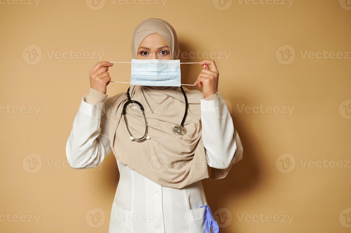 Muslim doctor with covered head in hijab holding and wearing medical mask standing on beige background eith copy space photo