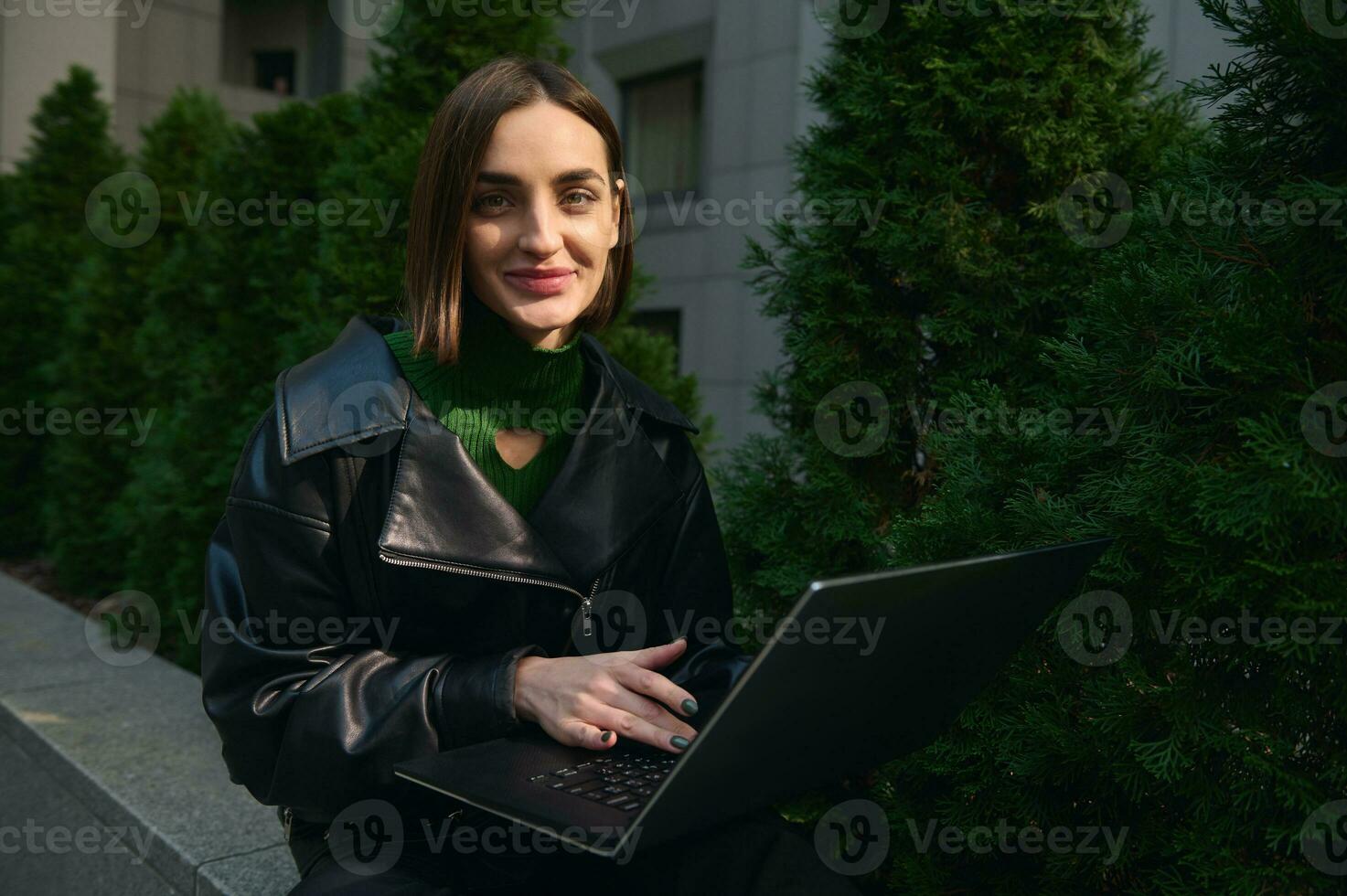 Close-up portrait of a young cute woman freelancer, copywriter, motivated start-up sitting on parapet with laptop, working remotely and smiling cute for camera. Connection and online business concept photo