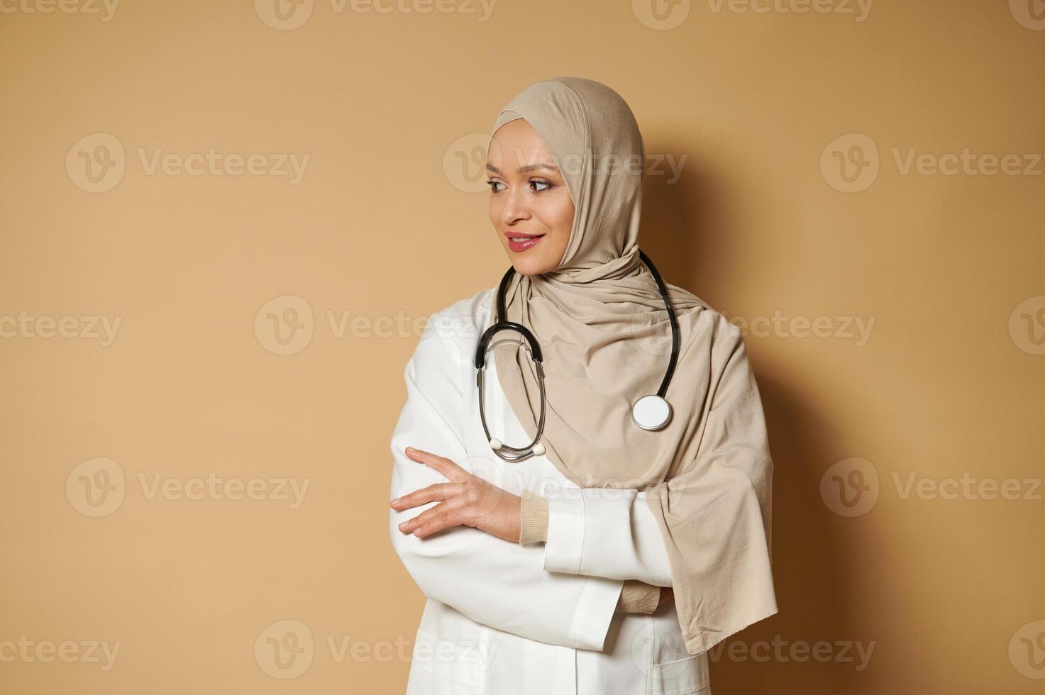 Beautiful serene Muslim doctor woman in hijab standing with crossed arms and looking side on beige background with copy space photo