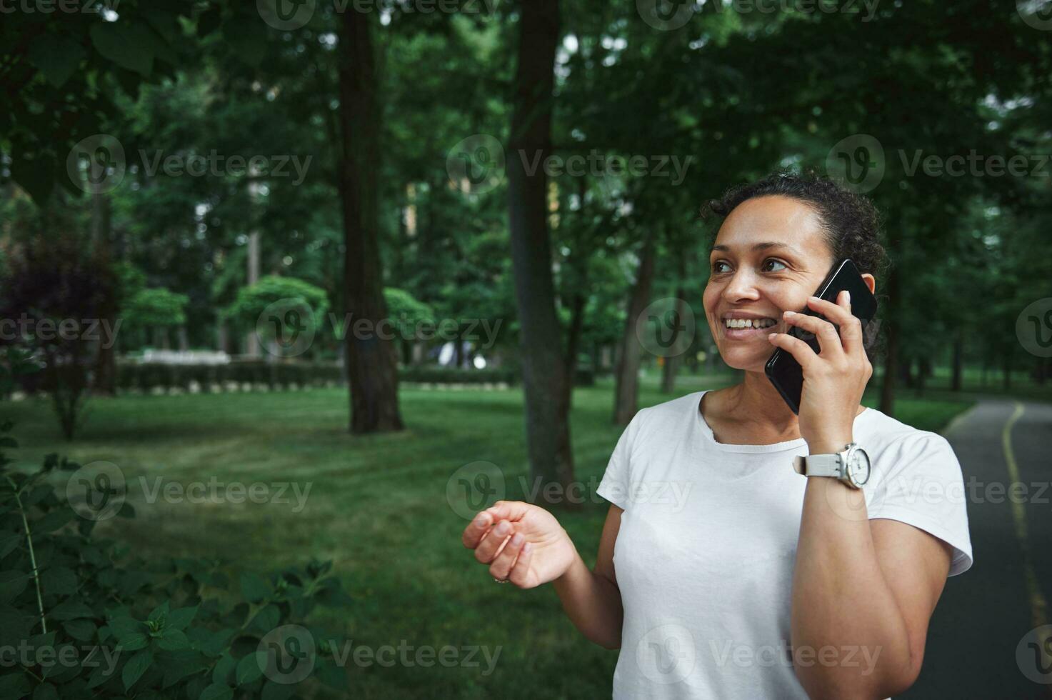 encantador multiétnico joven mujer hablando en móvil teléfono mientras vagante el callejón de un ciudad parque en un verano día foto