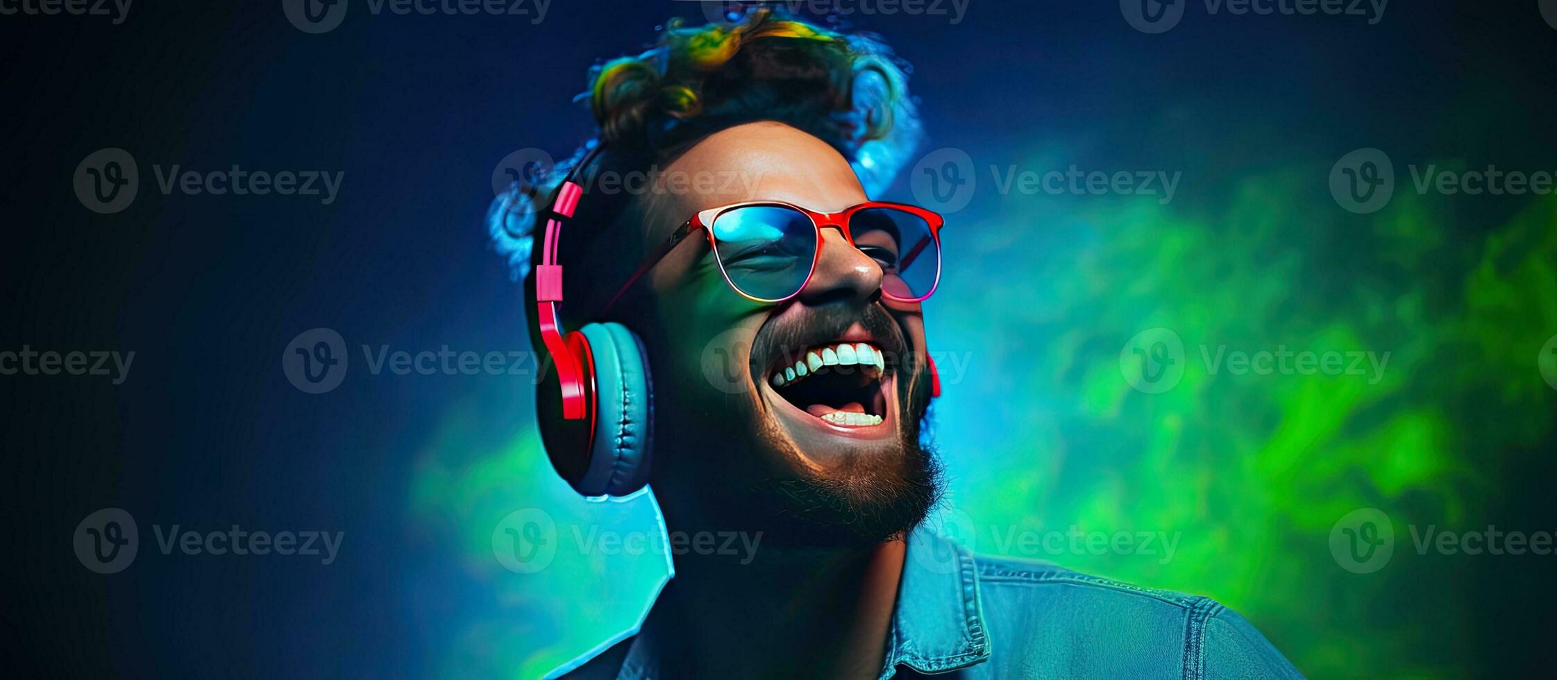Hipster man wearing headphones dancing and singing with an open mouth smile in a portrait with a blue background and mixed neon light showcasing his fashi photo