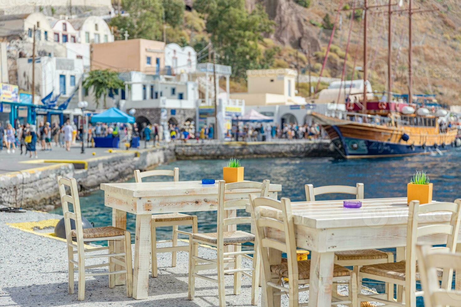 Chairs and tables of a traditional Greek taverna, restaurant at the dock of small port of blurred sail boat and tourist waking and shops with white architecture in Santorini Greece photo