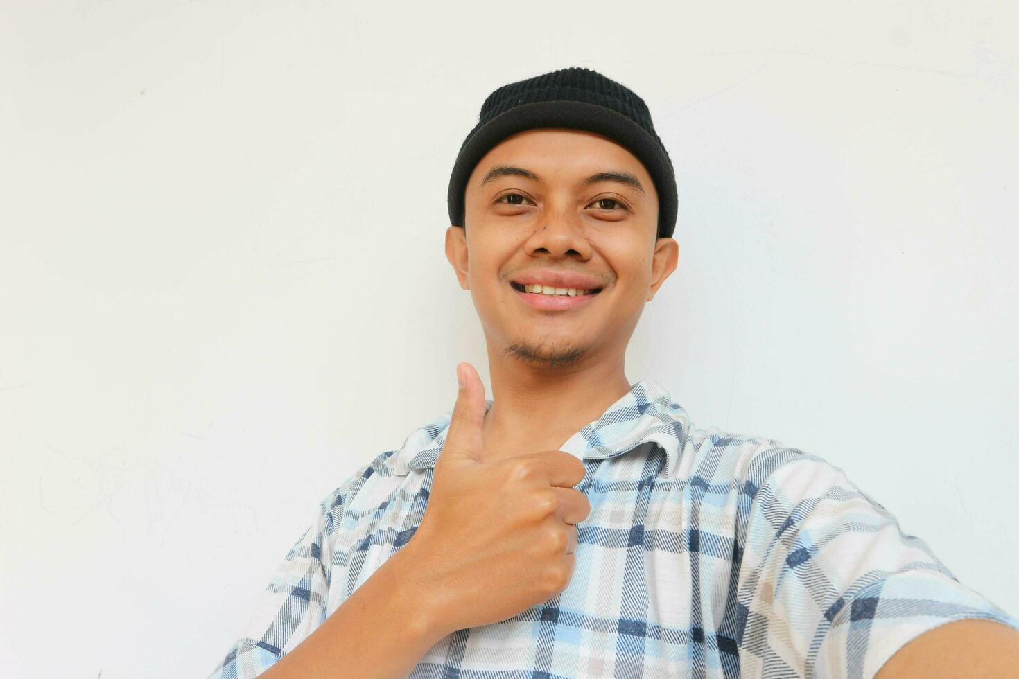 Happy adult man wearing pajamas and beanie cap smiling while showing thumb up and looking at camera, OK sign. Isolated on white background photo