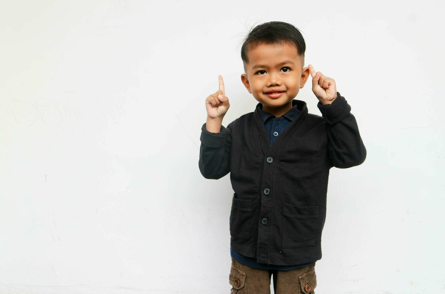 A smiling toddler boy looks at the camera raising his hands and pointing up, isolated on white background photo