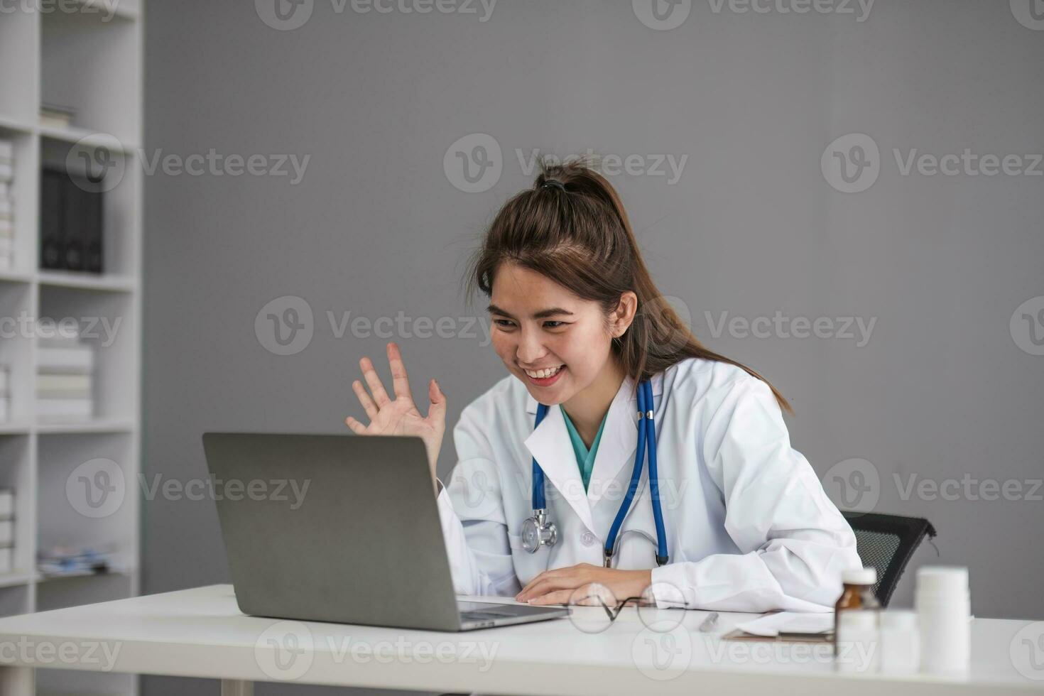 Young Asia lady doctor in white medical uniform with stethoscope using computer laptop talking video conference call with patient at desk in health clinic or hospital. Consulting and therapy concept. photo