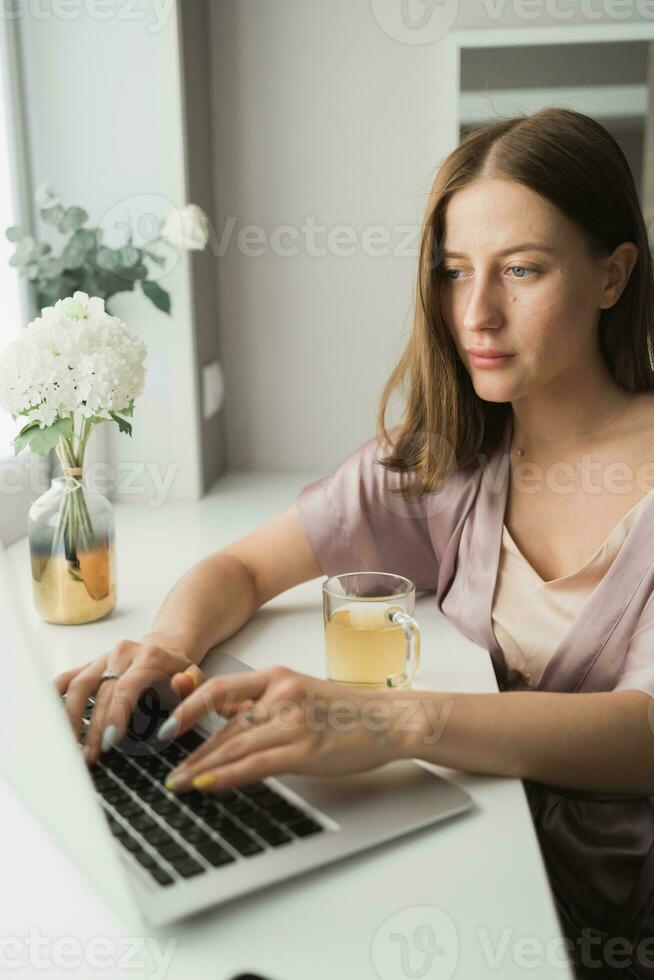 Young woman wears sleeping clothes working on laptop computer while sitting at living room and drinking tea - social network and working at home concept photo