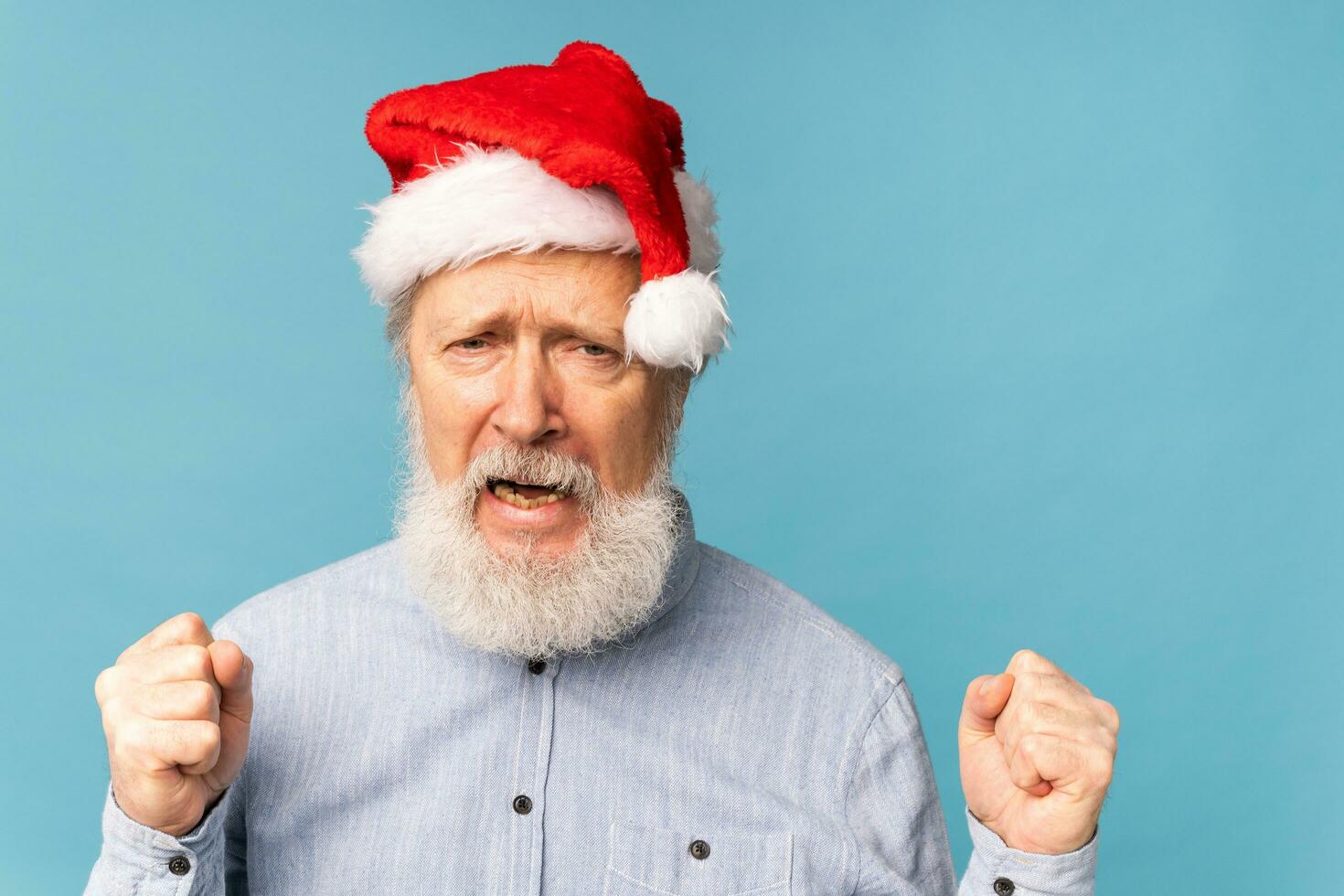 Happy confident cool old bearded Santa Claus winner raising fists celebrating triumph and success over blue background with copy space photo