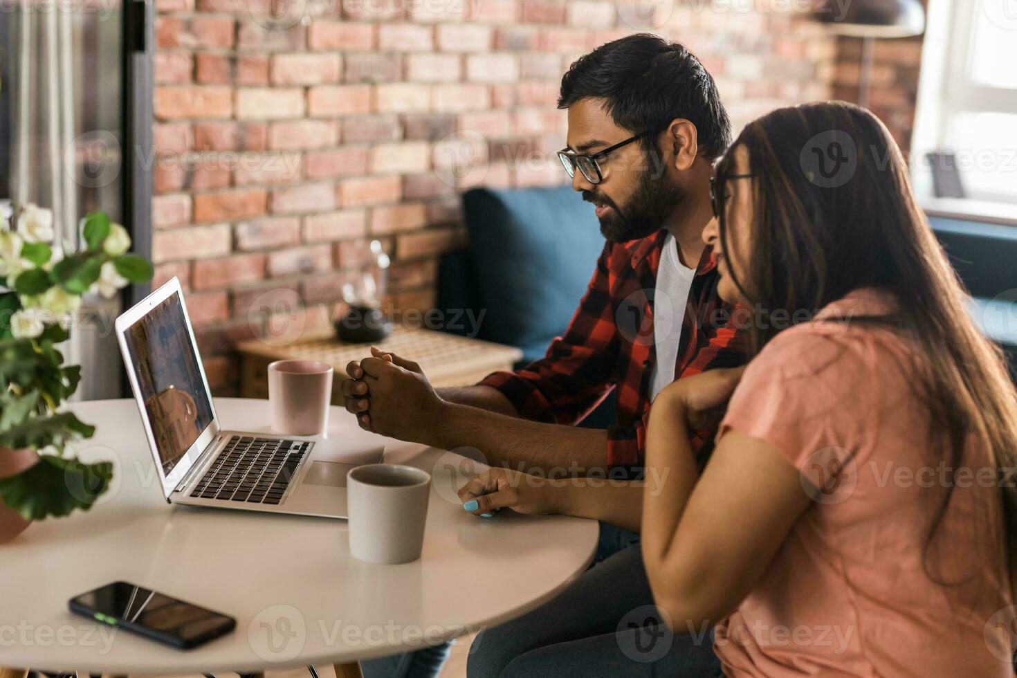 Indian family couple cuddle at desk make video call to friends using laptop webcam. Loving young spouses look at computer screen waving hands  communicating online via app photo
