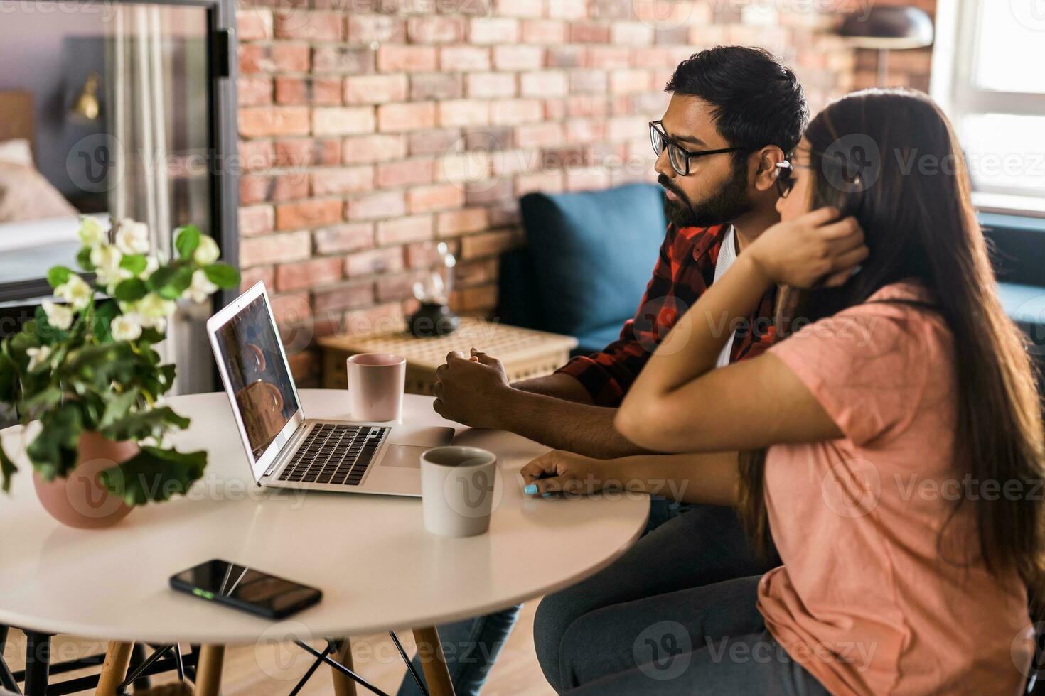 Indian family couple cuddle at desk make video call to friends using laptop webcam. Loving young spouses look at computer screen waving hands  communicating online via app photo
