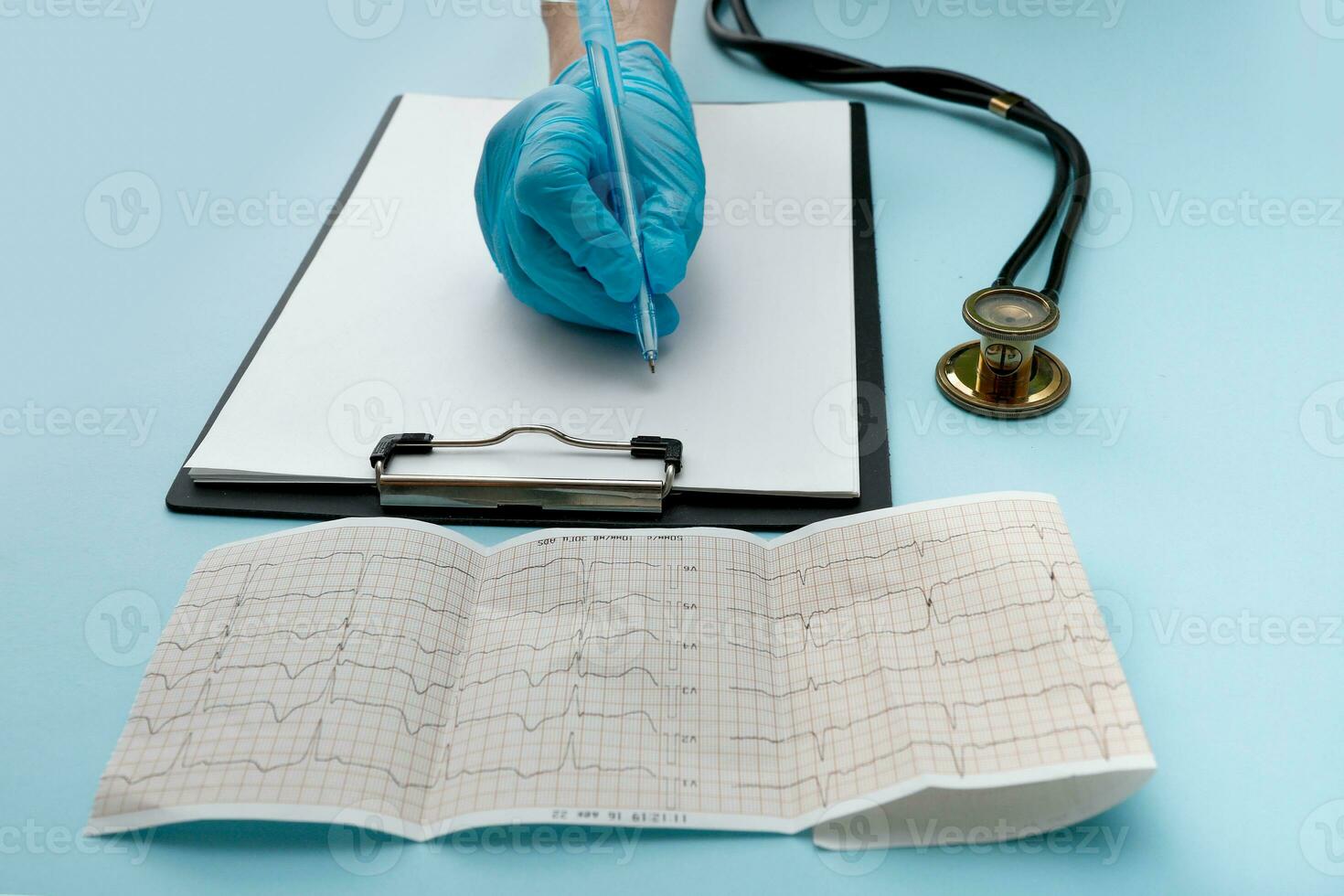 doctor's hand in a glove writes something on a tablet with a paper with a pen next to it lies a phonendoscope and a cardiogram photo