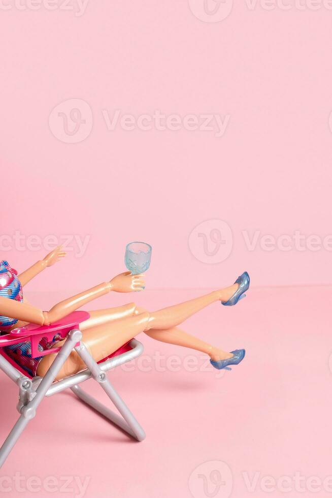 Doll with wineglass in beach chear on pink background. Beach relax concept. photo