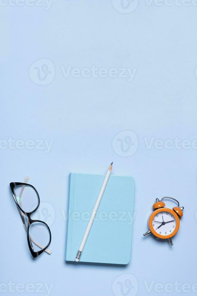 Notebook, glasses and alarm clock on blue background with copy space. Business minimalist concept photo